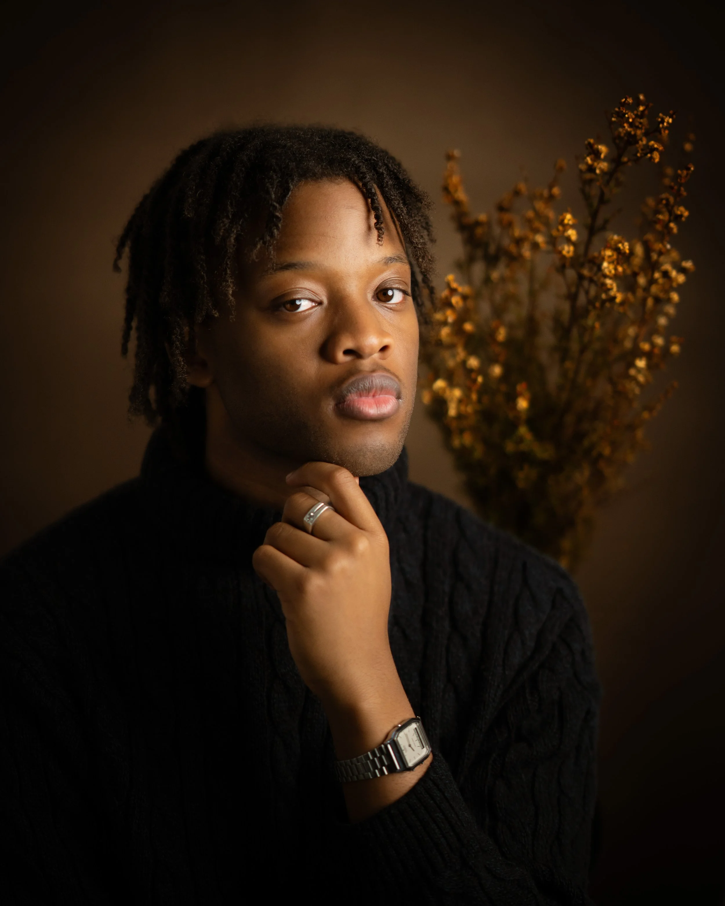 A young man with dreadlocks and brown eyes, wearing a black sweater and silver wristwatch, holding his chin with his right hand against a dark background with dried flowers.