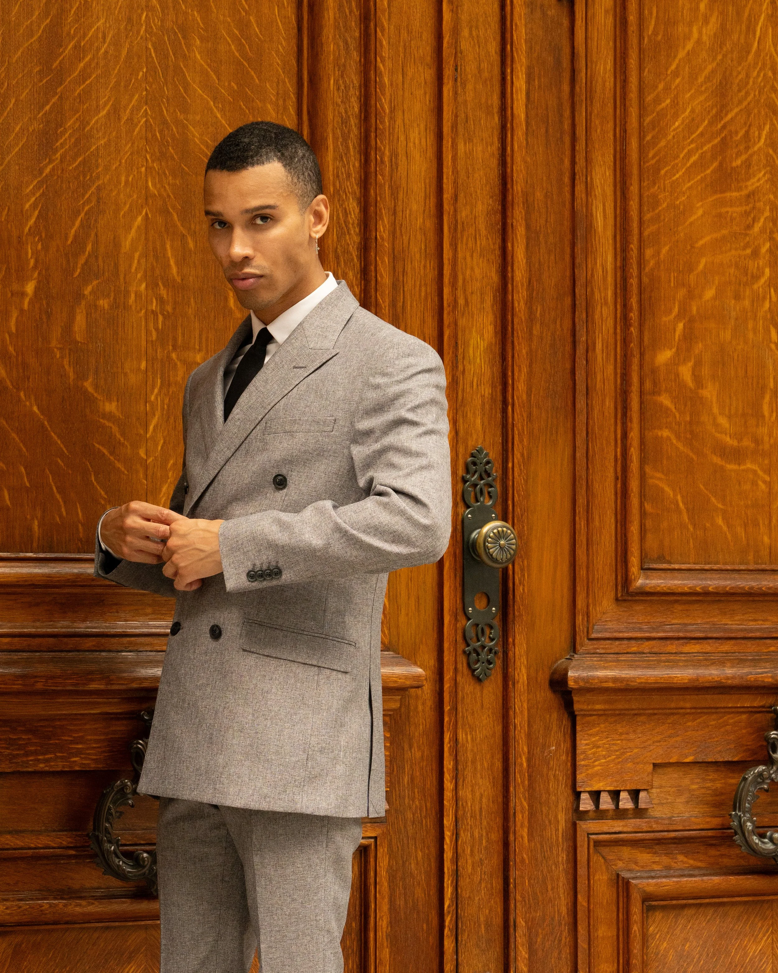 A man in a light gray suit with a white shirt and black tie standing in front of wooden doors with ornate handles