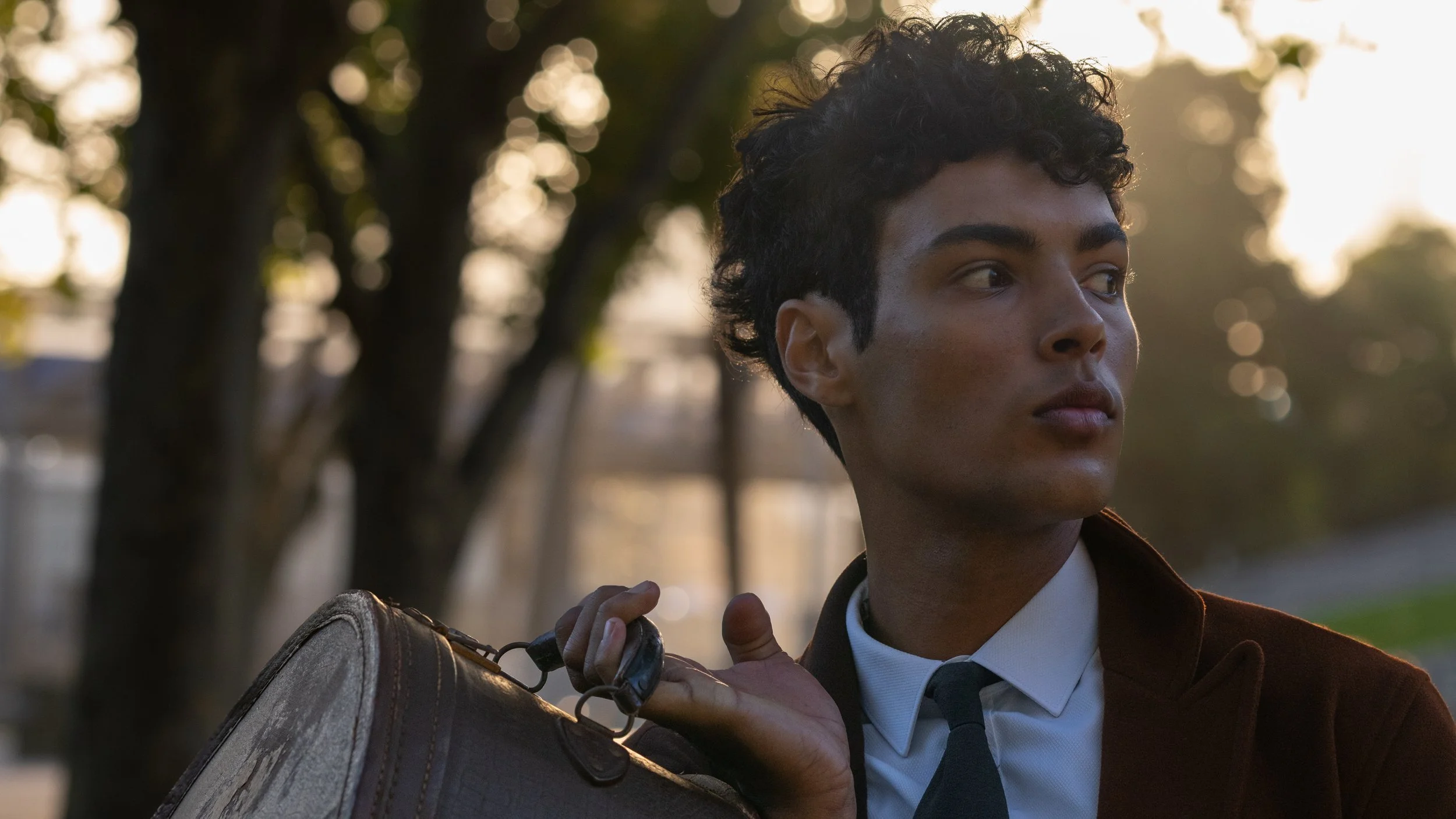 A young person with curly hair and medium skin tone stands outdoors during sunset, holding a guitar case over their shoulder, wearing a brown jacket over a white shirt and a dark tie.