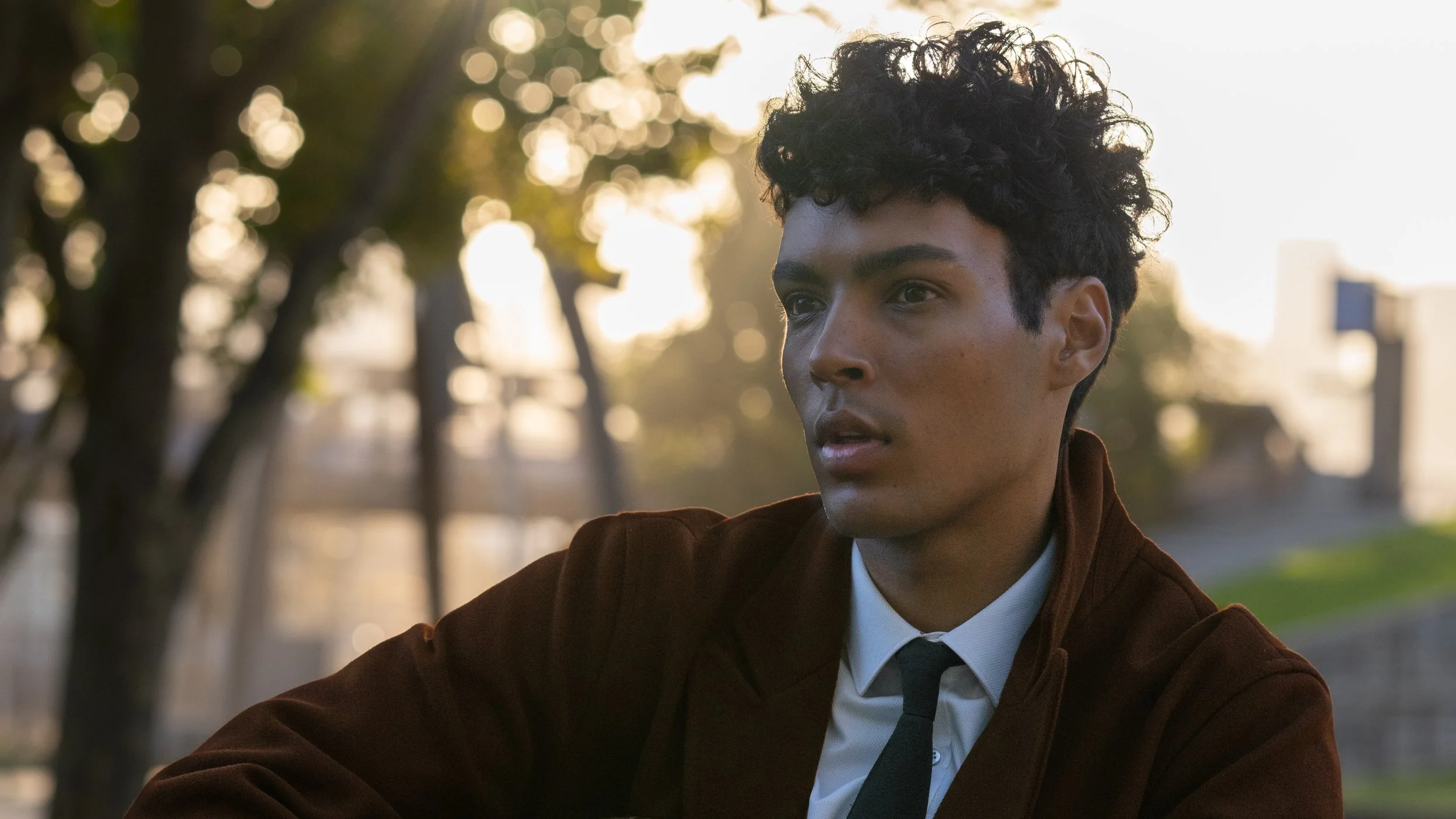 A young man with curly dark hair and a serious expression, wearing a brown jacket, white shirt, and a tie, sitting outdoors during sunset with trees and blurred city skyline in the background.