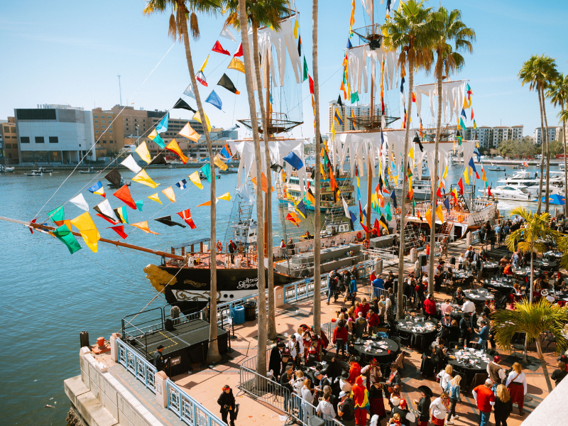 A lively outdoor event on a dock featuring a historic ship decorated with colorful flags and bunting, with people dining at tables under palm trees near a marina with boats and modern buildings in the background.