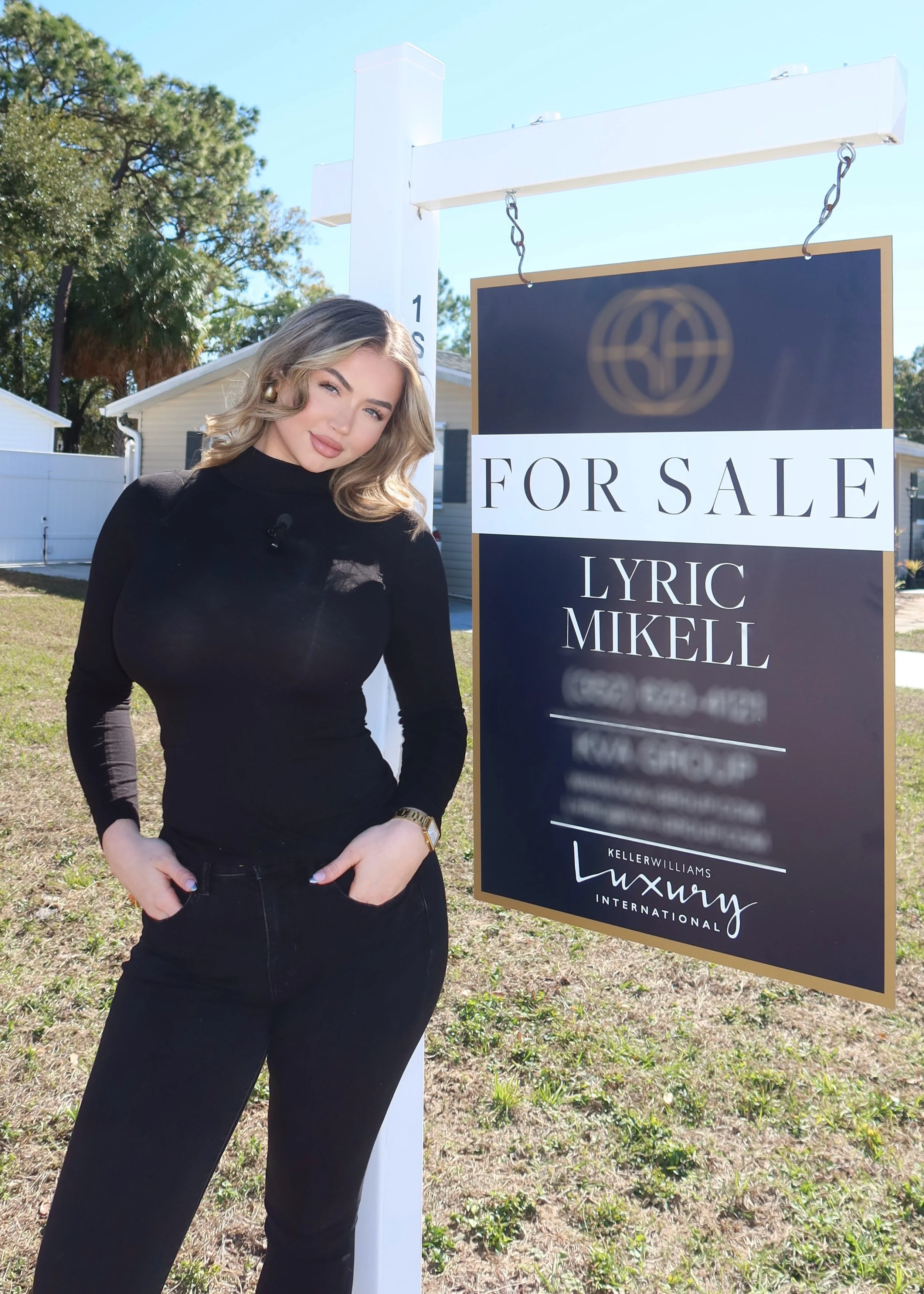 A woman with wavy blonde hair, wearing a black turtleneck and black pants, standing next to a for sale sign on a lawn with trees and houses in the background.