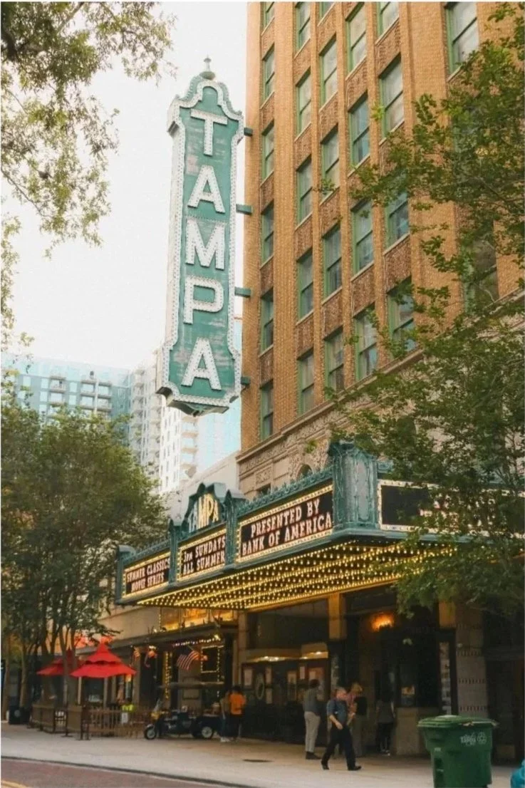 The historic Tampa theater marquee with a large vertical sign that reads 'TAMPA' in capital letters, situated on a city street with trees and pedestrians.