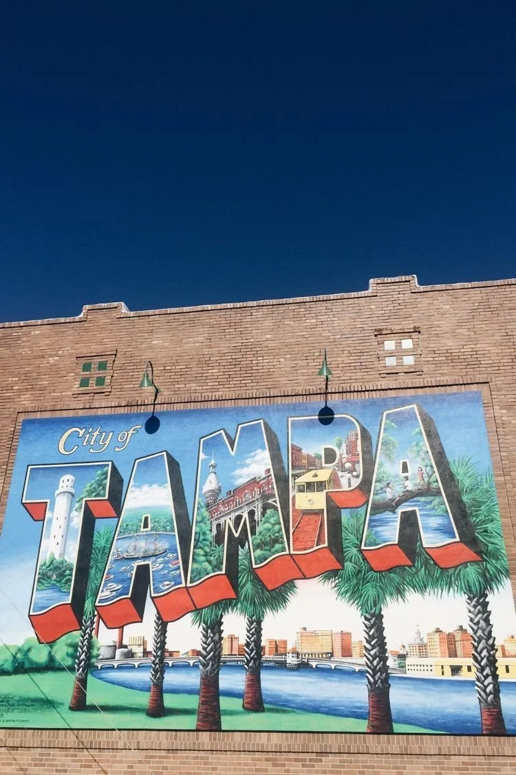Colorful mural on brick building spelling 'Welcome to TAMPA' with landmarks, palm trees, and cityscape in Tampa, Florida.