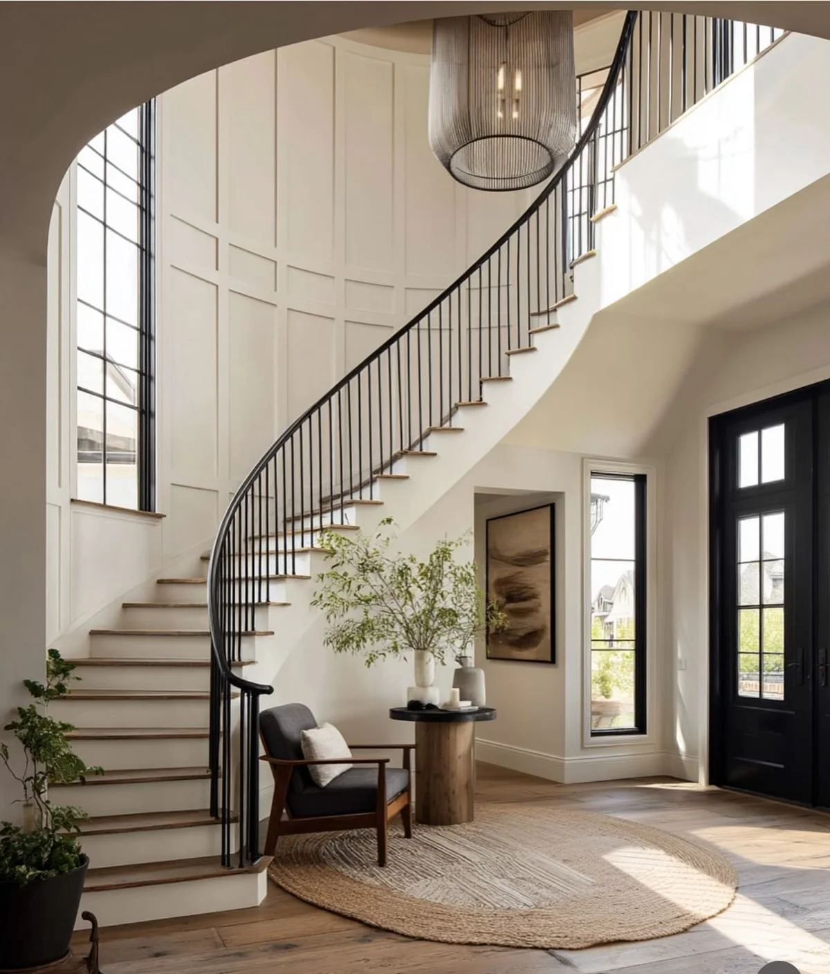 Interior view of a modern entryway with a curved staircase, black railing, large windows, a round rug, a dark front door, a armchair with a pillow, a small round wooden table with vases and a plant, and a framed artwork on the wall.