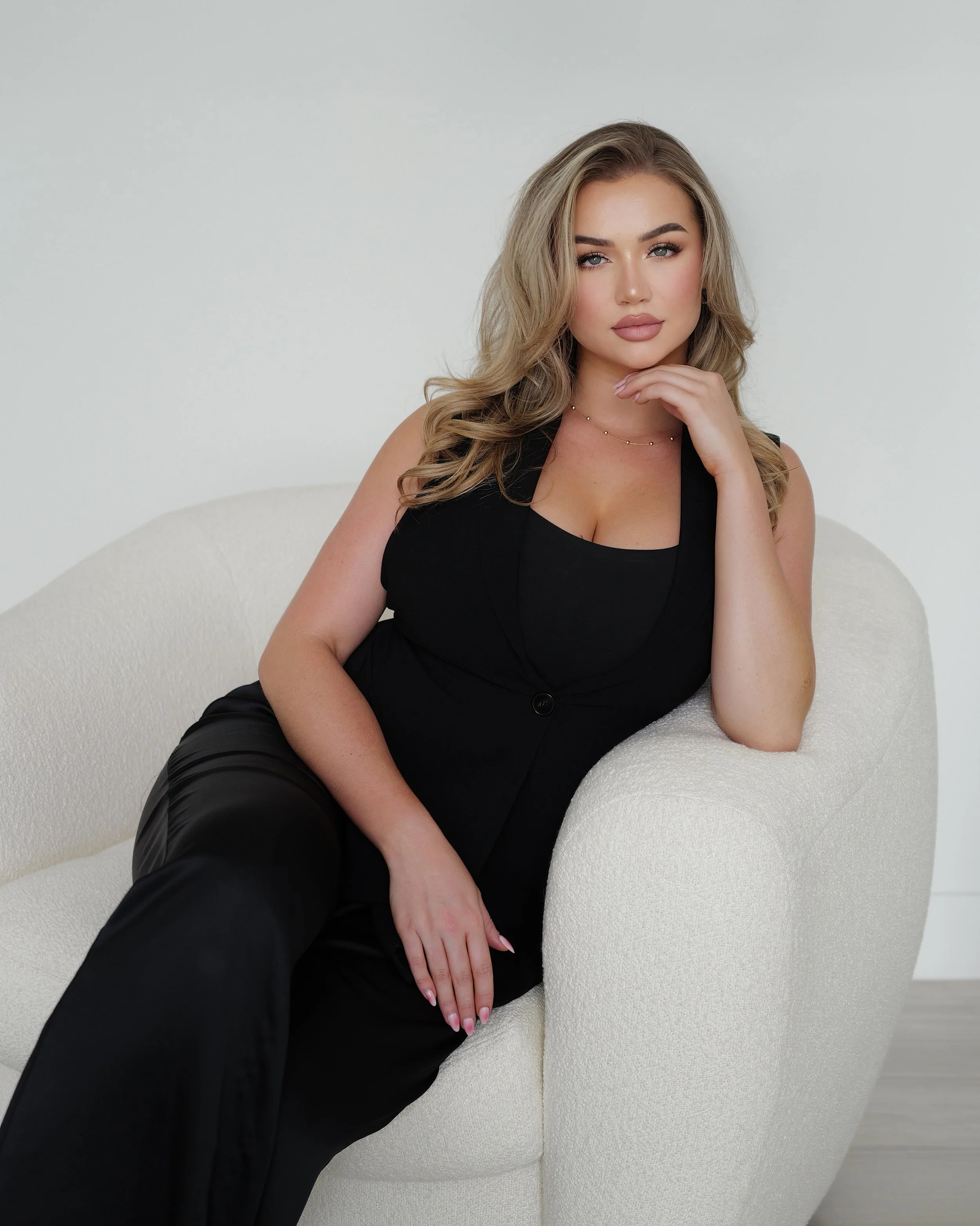 A woman with blonde, wavy hair seated on a light-colored couch in a black sleeveless outfit, looking at the camera with her right hand resting on her chin.