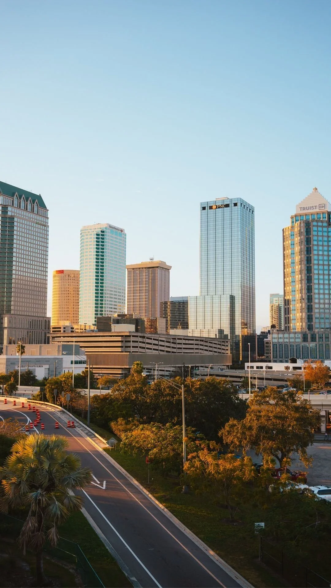 City skyline with tall glass office buildings, trees lining the streets, and a clear blue sky.