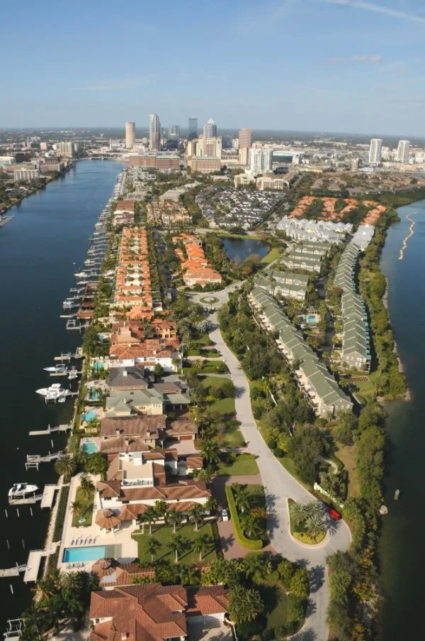 Aerial view of a city with high-rise buildings in the background, water canals on both sides, and residential houses with pools in the foreground.