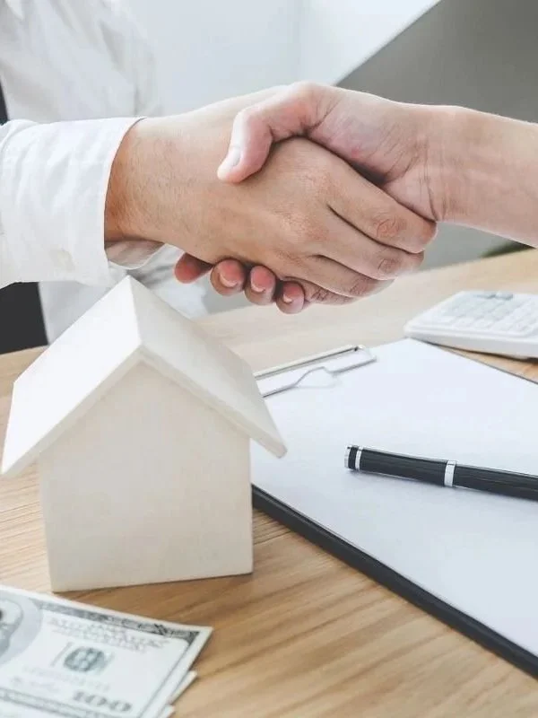 Two people shaking hands over a table with a small model house, a clipboard, a pen, and some hundred-dollar bills.