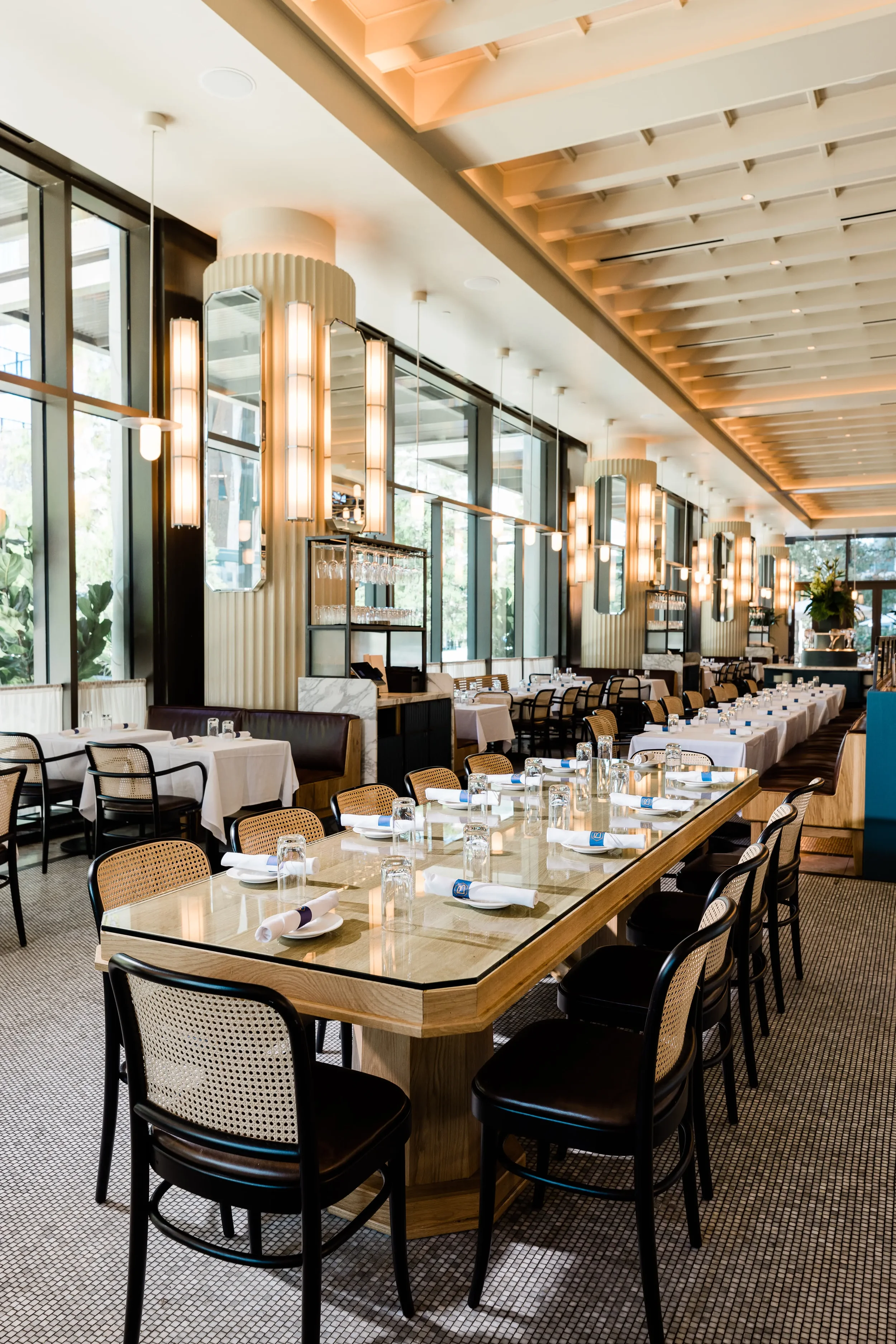 Elegant restaurant interior with a long wooden dining table set with white napkins and glasses, surrounded by black and cane chairs, featuring tall lighting fixtures and large windows letting in natural light.