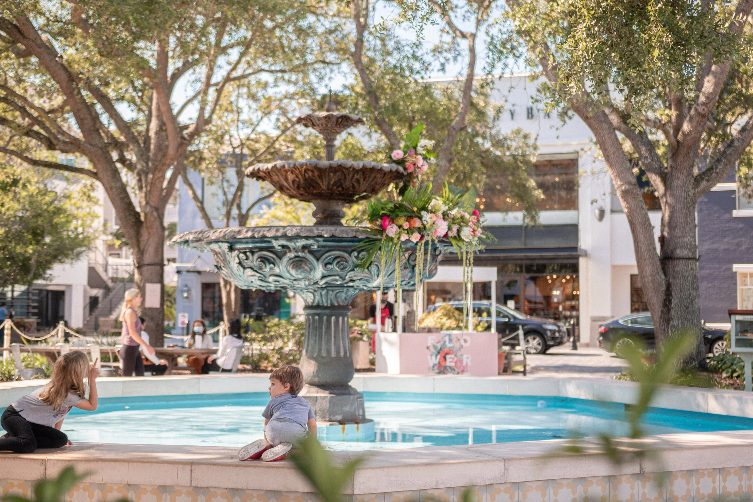Children playing by a decorative fountain in an outdoor plaza with trees and shops in the background.