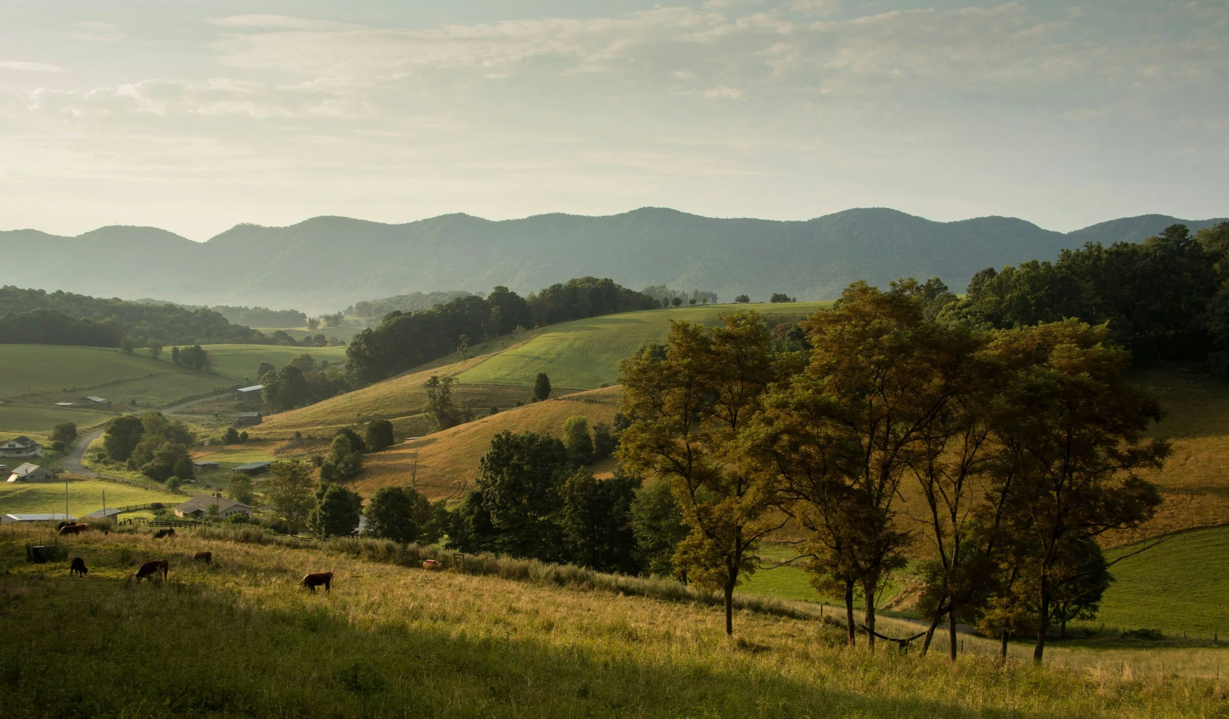 Scenic view of rolling green hills and mountains with trees and a small farm, under a partly cloudy sky.