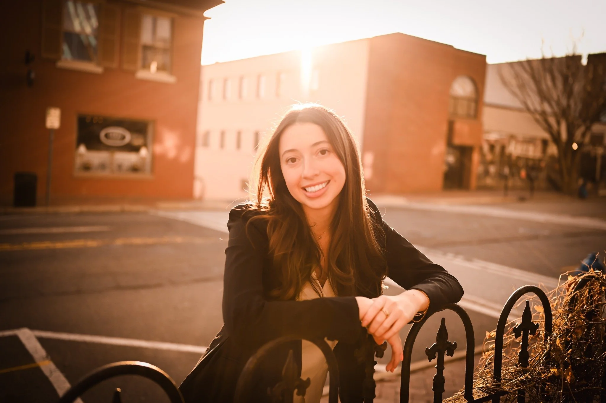 A young woman with long brown hair smiling, leaning on a black metal fence on a city street at sunset.