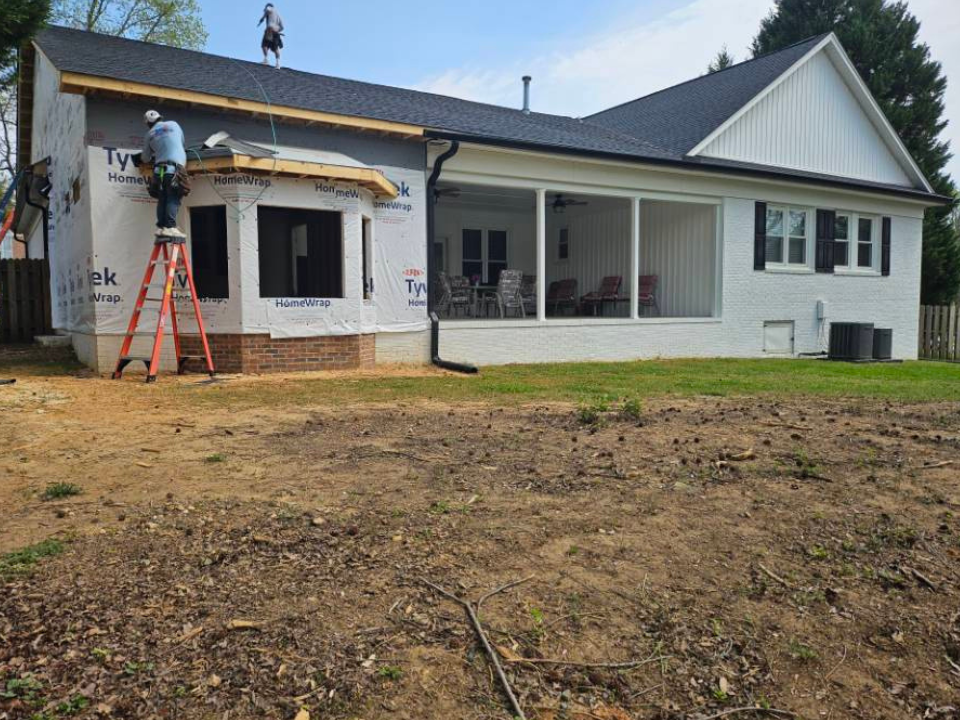 Construction workers working on the exterior of a house, with one on a ladder and another on the roof, installing or repairing the roof structure. The house is partially finished with some siding and brickwork visible.