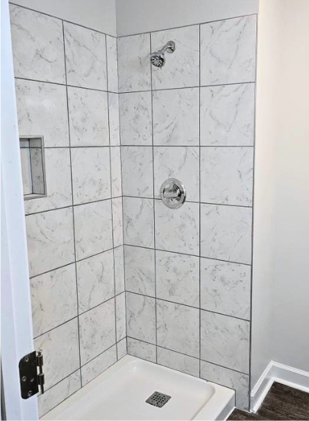 Empty shower with white marble tile walls, a chrome showerhead, and a chrome control handle, inside a bathroom with gray walls and dark wood flooring.