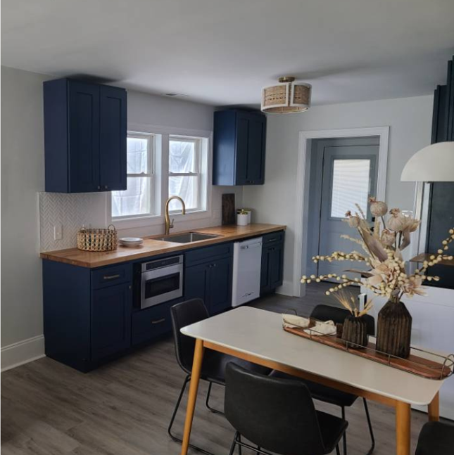 Kitchen with blue cabinets, white walls, wooden countertop, and a dining table with black chairs and a floral centerpiece.