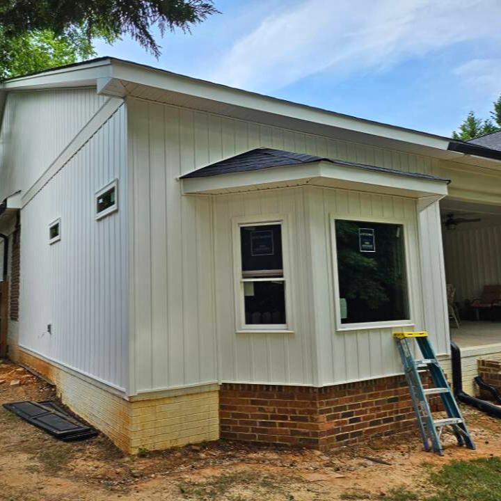 Newly installed white siding on the exterior of a house with a brick foundation, with a small porch area and a ladder leaning against it.