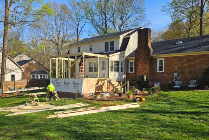 Construction of a wooden deck addition to the back of a house with workers and building materials in the yard.