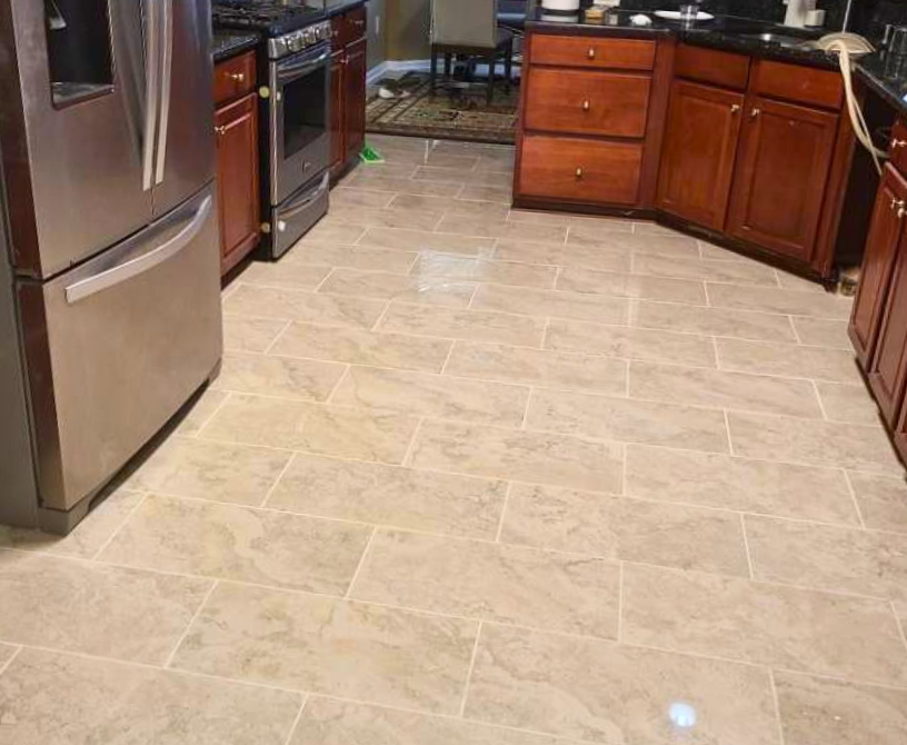 A kitchen with beige tile flooring, stainless steel refrigerator, dark wood cabinets, and a dining area in the background.