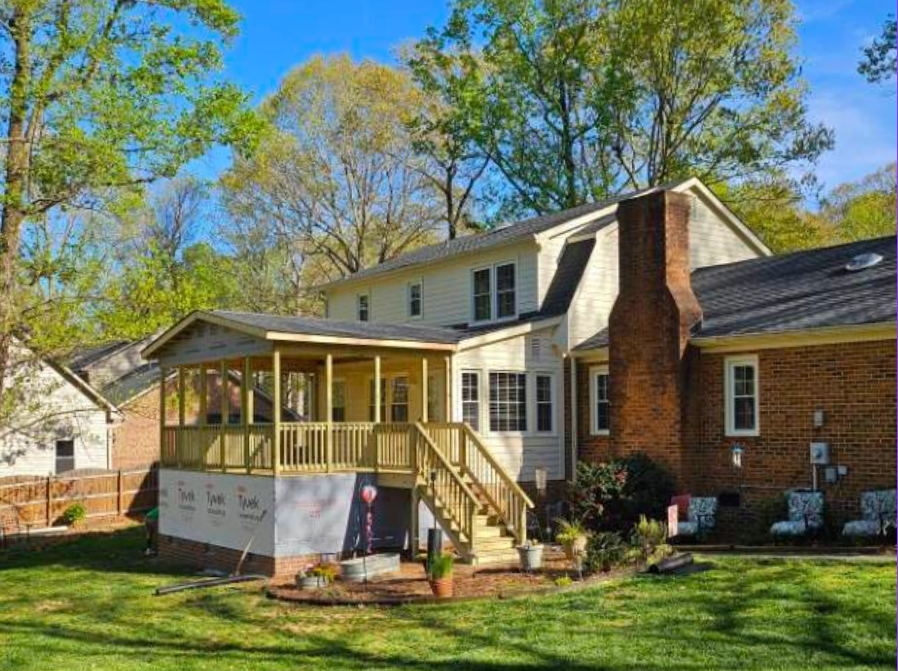 A two-story house with a brick chimney and a porch under construction, surrounded by a lawn with outdoor chairs and trees with green leaves.