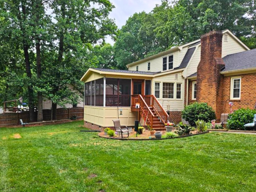 A backyard with a well-maintained lawn, surrounding tall trees, and a two-story house with a screened porch, brick chimney, and sitting area with chairs and potted plants.