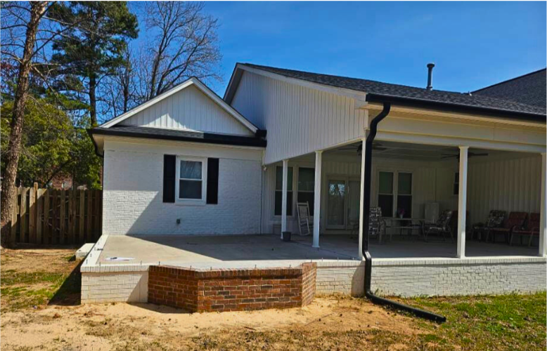 Backyard view of a white house with a covered porch, brick and concrete patio, black gutter, and a wooden fence on the left, under a clear blue sky.