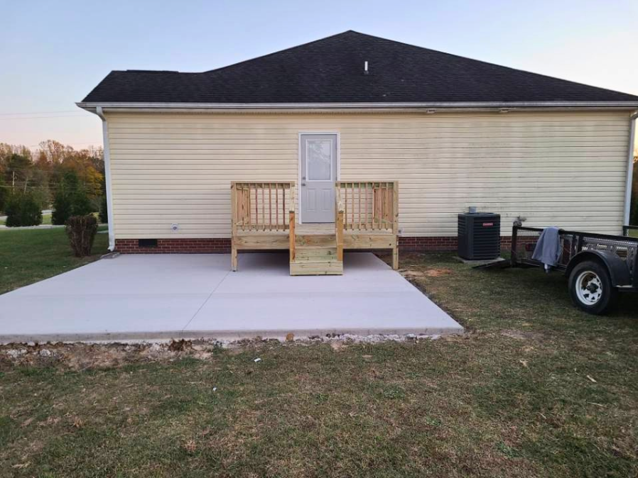 A small wooden deck with stairs and railing attached to the back of a house. The house has beige siding and a dark shingled roof. There is a concrete patio and a grassy yard with some equipment on the side.