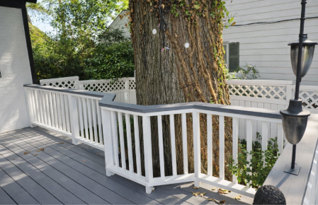 A porch with white railing and a large tree trunk in the center, with a black lantern-style outdoor light fixture attached to the railing, and trees and house in the background.