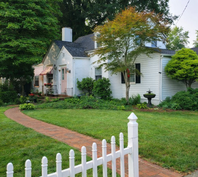 White house with a black roof, surrounded by green trees and bushes, with a brick pathway leading to the front porch, a white picket fence in the foreground, and a manicured lawn.