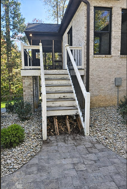 The image shows outdoor stairs with a porch at the top, attached to a house with beige brick walls. The stairs appear weathered and damaged at the bottom, exposing the underlying support structure. The area around the stairs is landscaped with small 