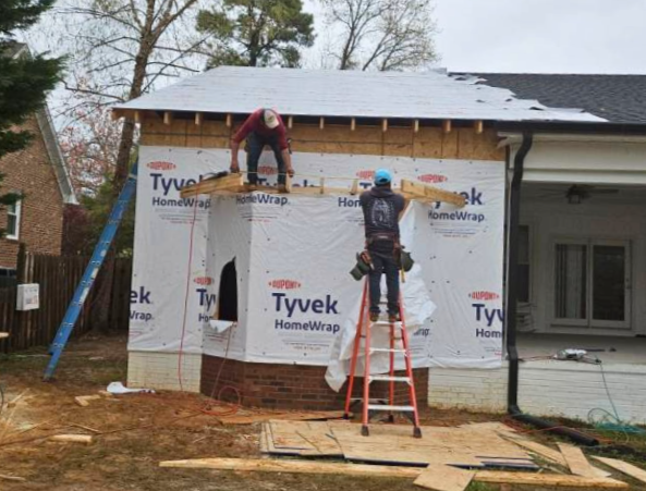 Two construction workers installing house wrap on a small building with a sloped roof, using ladders and tools, with trees and neighboring houses in the background.