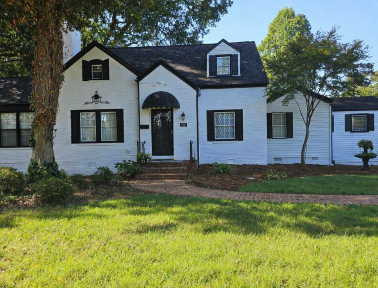 White house with black trim, brick stairs, and a curved awning over the front door, surrounded by green grass and trees.