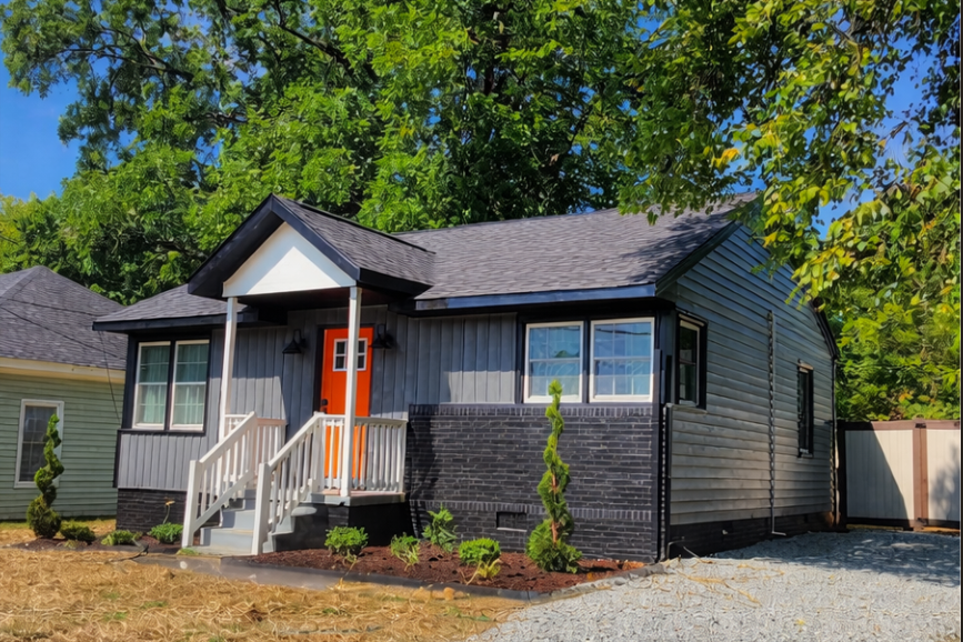 A newly renovated small house painted in gray and black with an orange front door, white stairs, surrounded by trees and mulch, with neighboring houses and a gravel driveway.