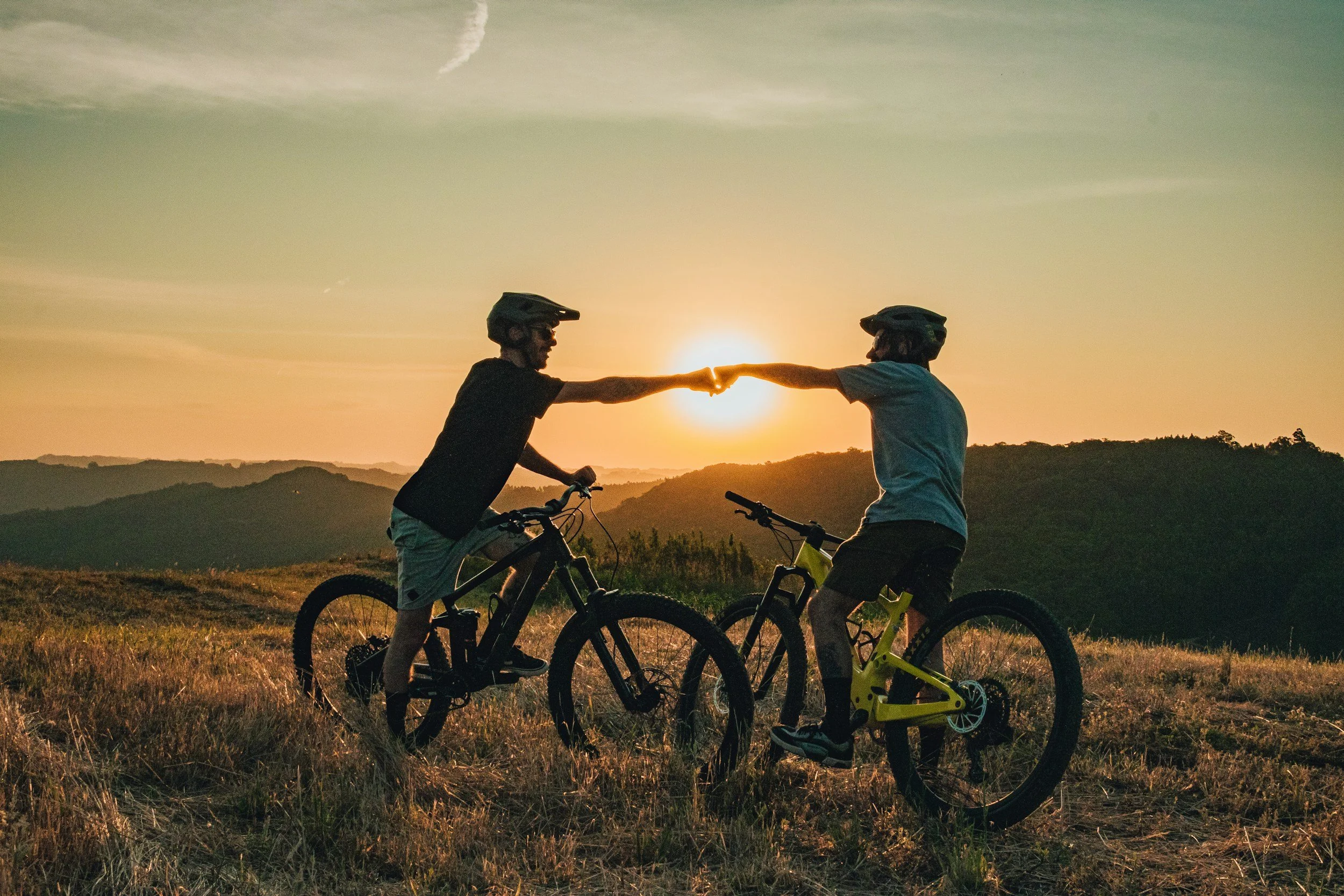 Two people with mountain bikes exchanging a fist bump at sunset in a scenic outdoor area with hills in the background.