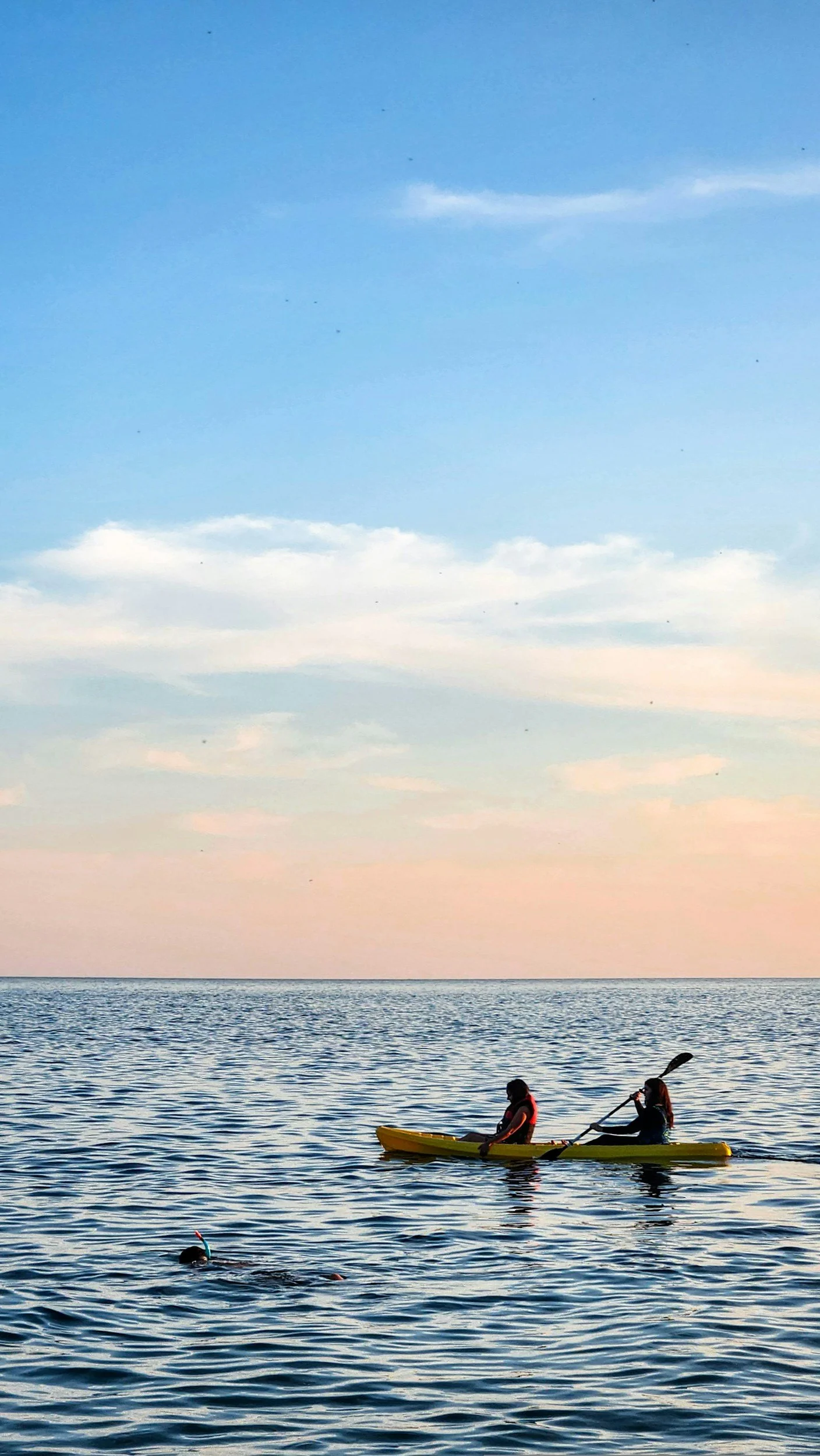Two people kayaking in a body of water during sunset or sunrise with a pastel-colored sky and some clouds.