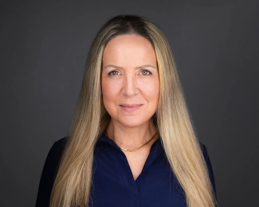 Professional portrait of a woman with long blonde hair wearing a navy blue blouse and a gold chain necklace, set against a gray background.