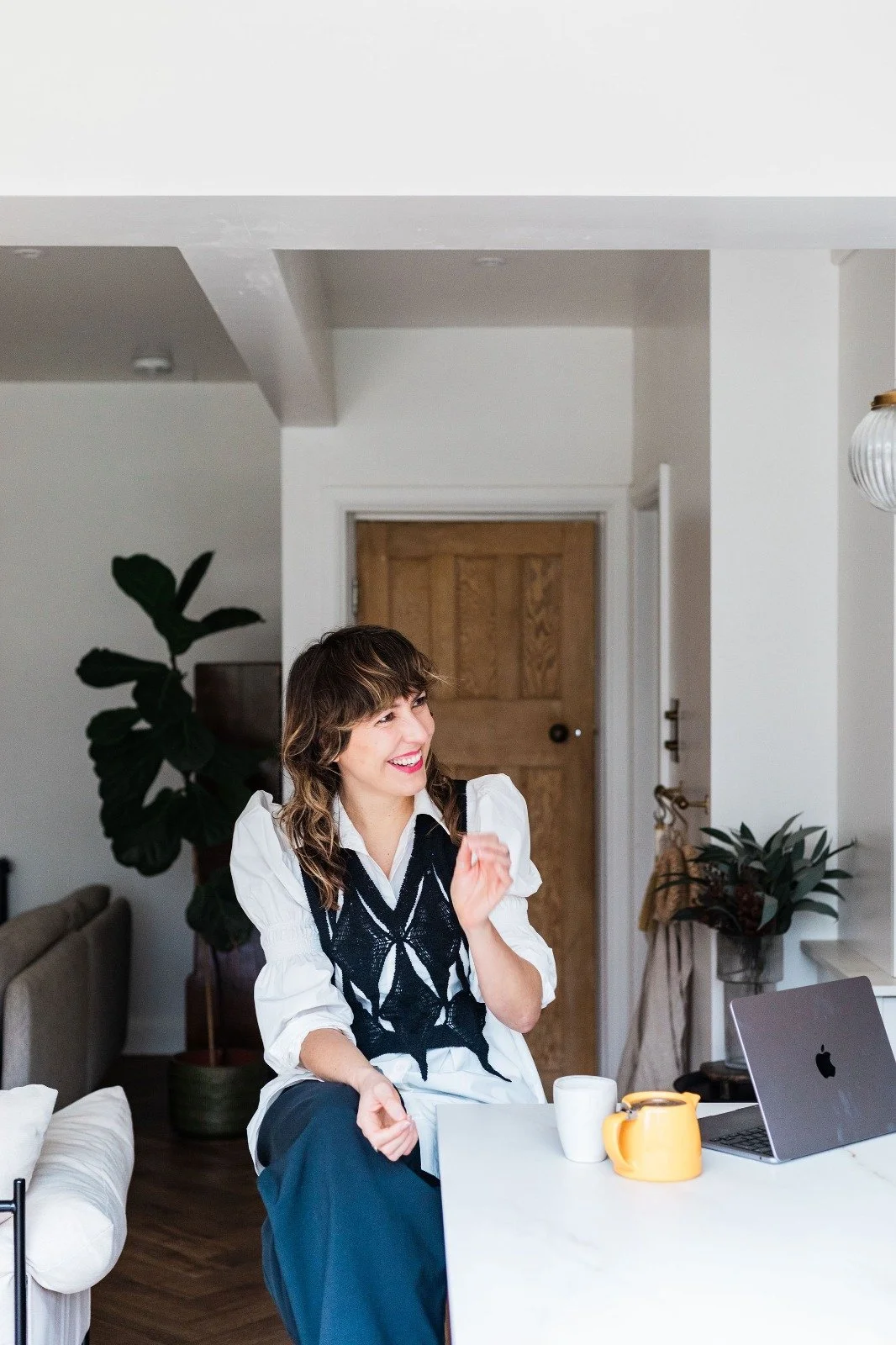 Becs Pearson copywriter sitting at a kitchen counter laughing with a laptop and yellow teapot in front of her