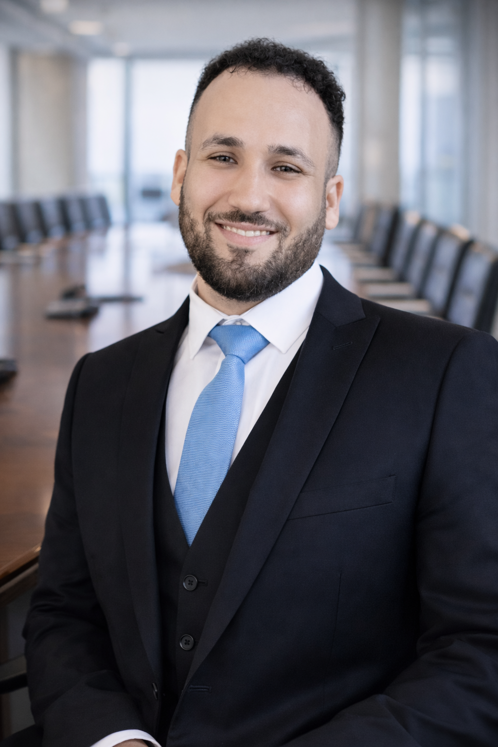A young man in a black suit, white shirt, and light blue tie smiling in a modern conference room.