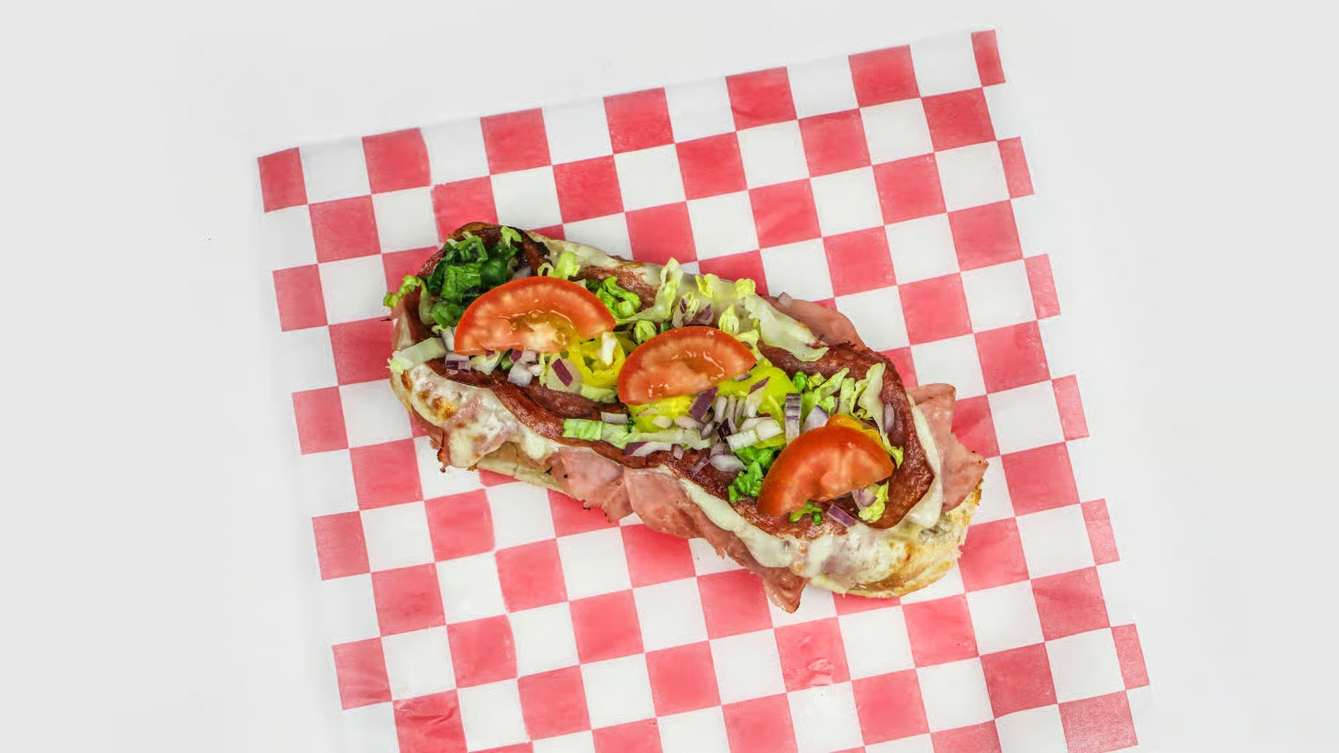 A hot dog topped with chopped lettuce, tomato slices, shredded cheese, and chopped onions, placed on a red and white checkered paper.