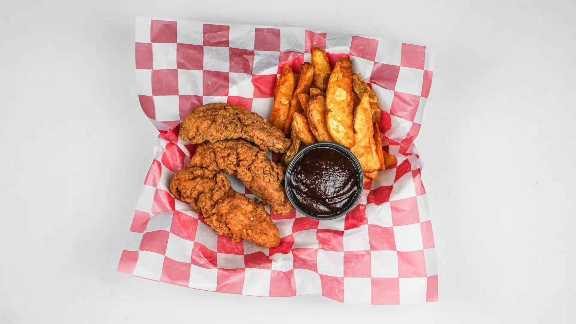 Fried chicken tenders, seasoned potato wedges, and a cup of barbecue sauce on checkered paper