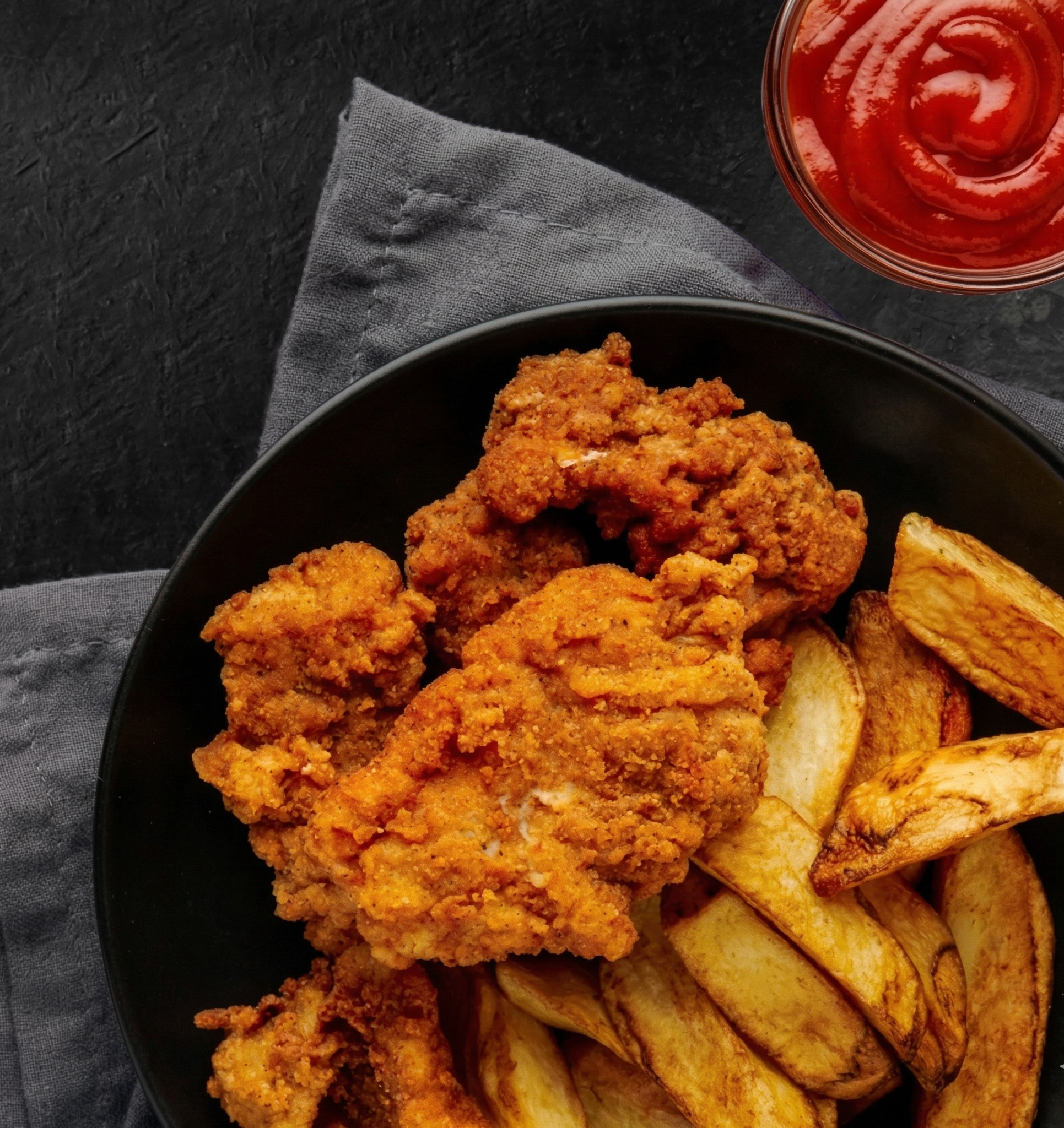 Fried chicken pieces with potato wedges served on a black plate, with a side of ketchup in a small glass bowl, on a dark textured surface with a gray cloth napkin.