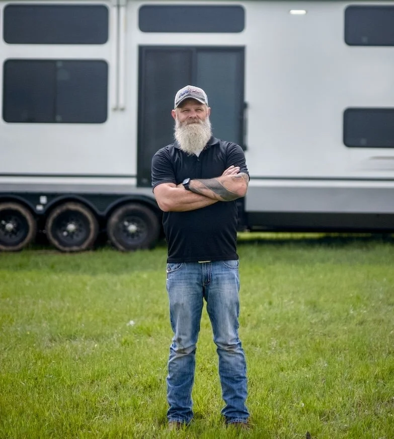 A man with a beard, wearing a baseball cap, black polo shirt, and jeans, standing with arms crossed in front of a large white trailer on a grassy field.