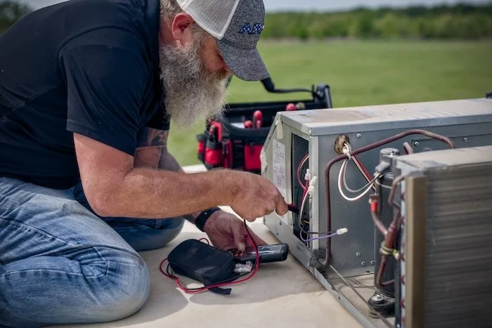 A man with a gray beard and gray cap kneels next to an open air conditioning unit, using a tool to perform maintenance or repairs.