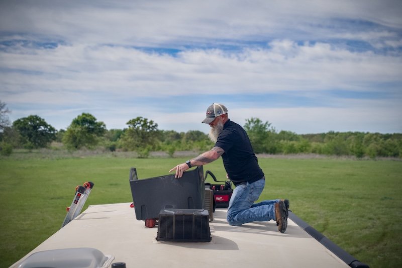 A man with a beard wearing a baseball cap, black t-shirt, and jeans kneeling on top of an RV, organizing tools and equipment for outdoor work or maintenance in a grassy field under a partly cloudy sky.