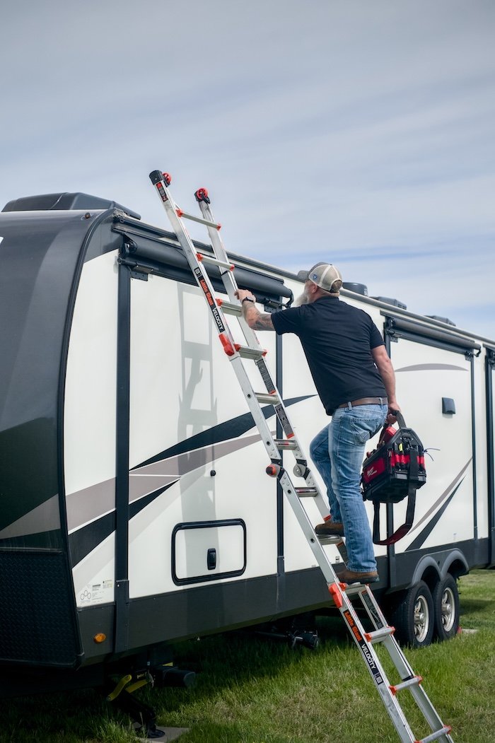 A man standing on a ladder inspecting the roof of a travel trailer, holding a toolkit in one hand, under a cloudy sky.