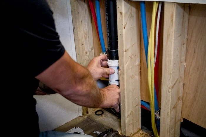 A person working on installing or repairing plumbing pipes in a wall with exposed wooden studs. The pipes are color-coded with blue, red, yellow, and black.