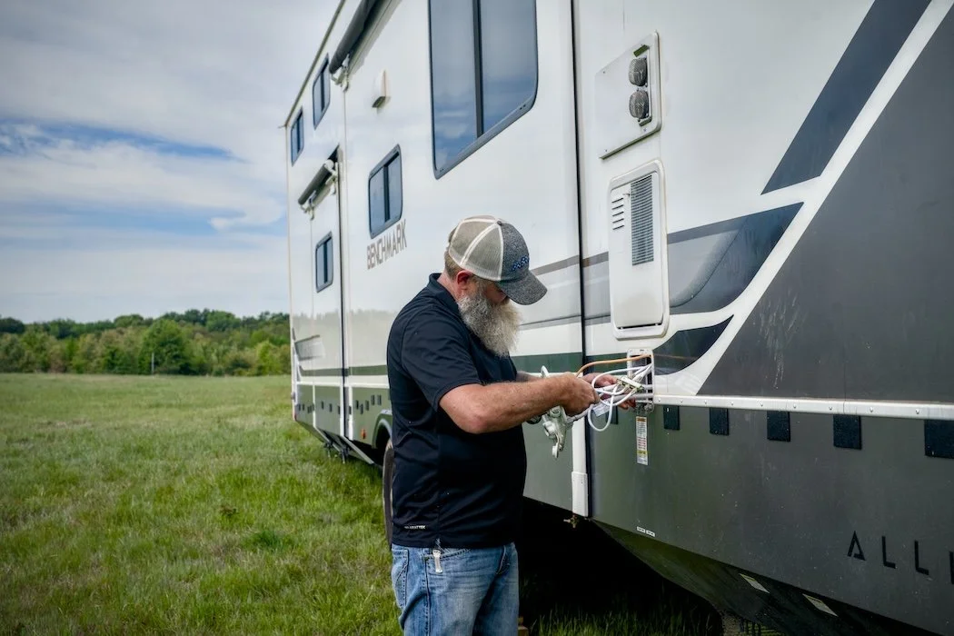 A man with a beard, wearing a baseball cap and black shirt, working on the exterior of a large recreational vehicle in a grassy field.