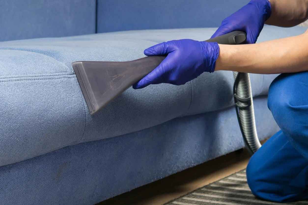 Person in blue gloves vacuuming a light blue sofa with a handheld vacuum cleaner.