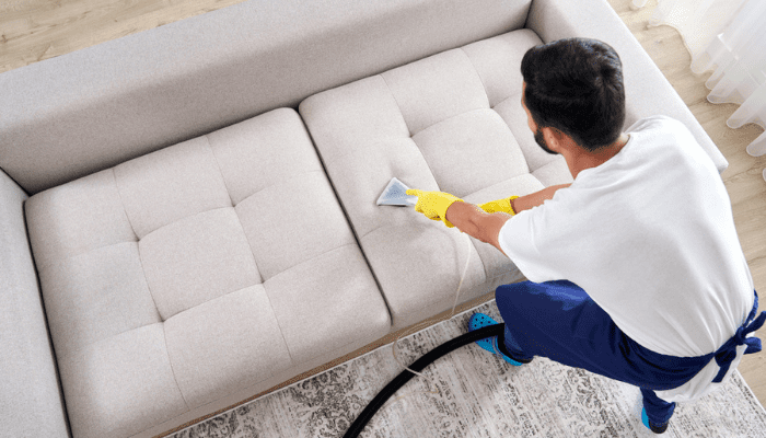 Man wearing yellow gloves cleaning beige sofa with handheld vacuum in living room.