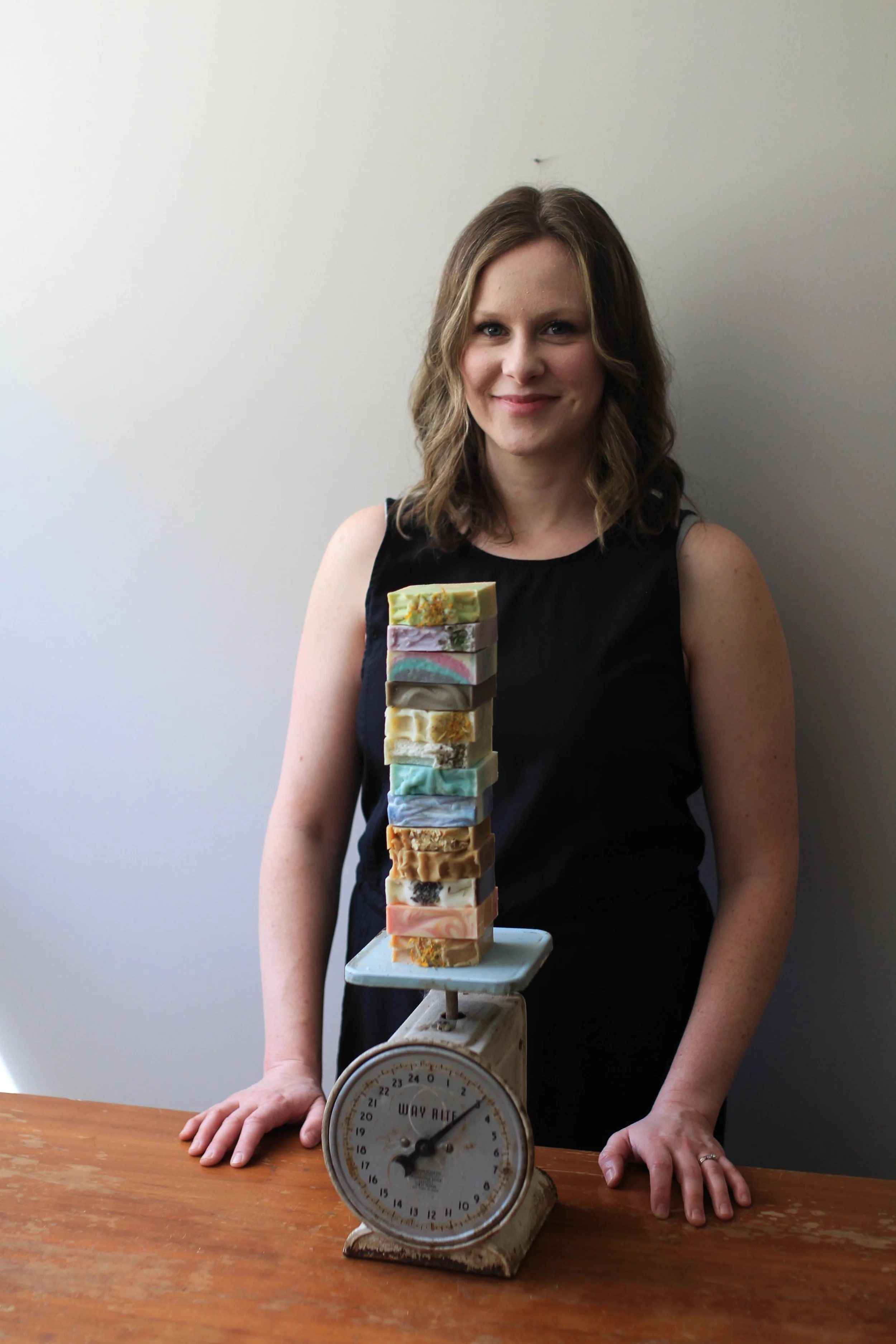 A woman with shoulder-length brown hair in a black sleeveless top stands behind a wooden table, smiling, with her hands resting on the table, next to a vintage kitchen scale. Stacked on the scale are various colorful handmade soap bars, arranged in a tall column, with a plain white background.
